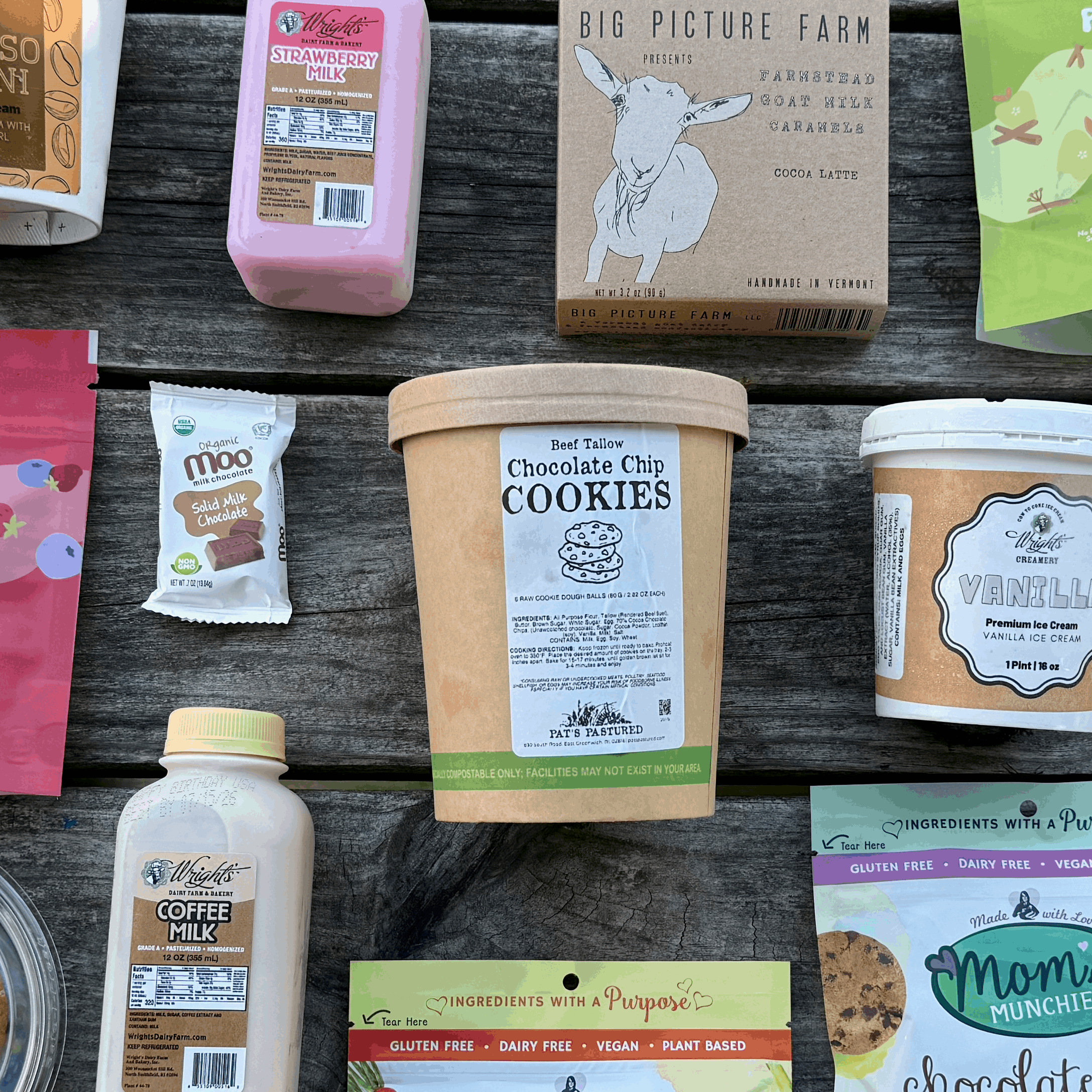 A collection of various snack, dessert, and coffee products arranged neatly on a weathered gray wooden picnic table outdoors. The products include Piquadits, Mom's, Picture Farm, and Exotic Farms items such as cookies, carrot bites, almond butter, apple bites, and cold drinks in plastic cups.