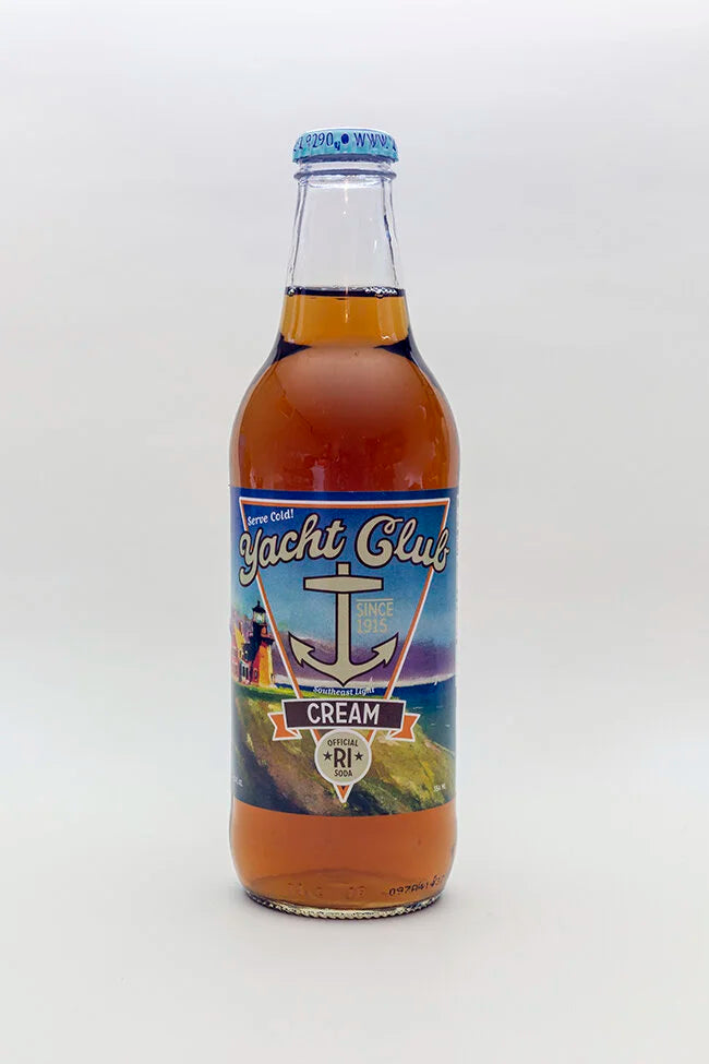 A bottle of Yacht Club Cream soda with amber liquid, a blue cap, and a blue and yellow label featuring an anchor and lighthouse, set against a plain light gray background.