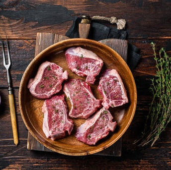 Raw lamb loin chops on a wooden cutting board with a sprig of thyme and a slice of onion beside them, on a rustic wooden table.